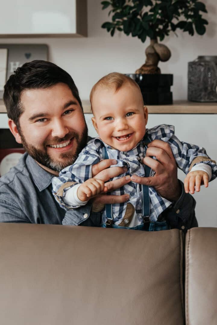 Ein Vater lacht mit seinem Sohn der er hinter dem Sofa hochhebt.