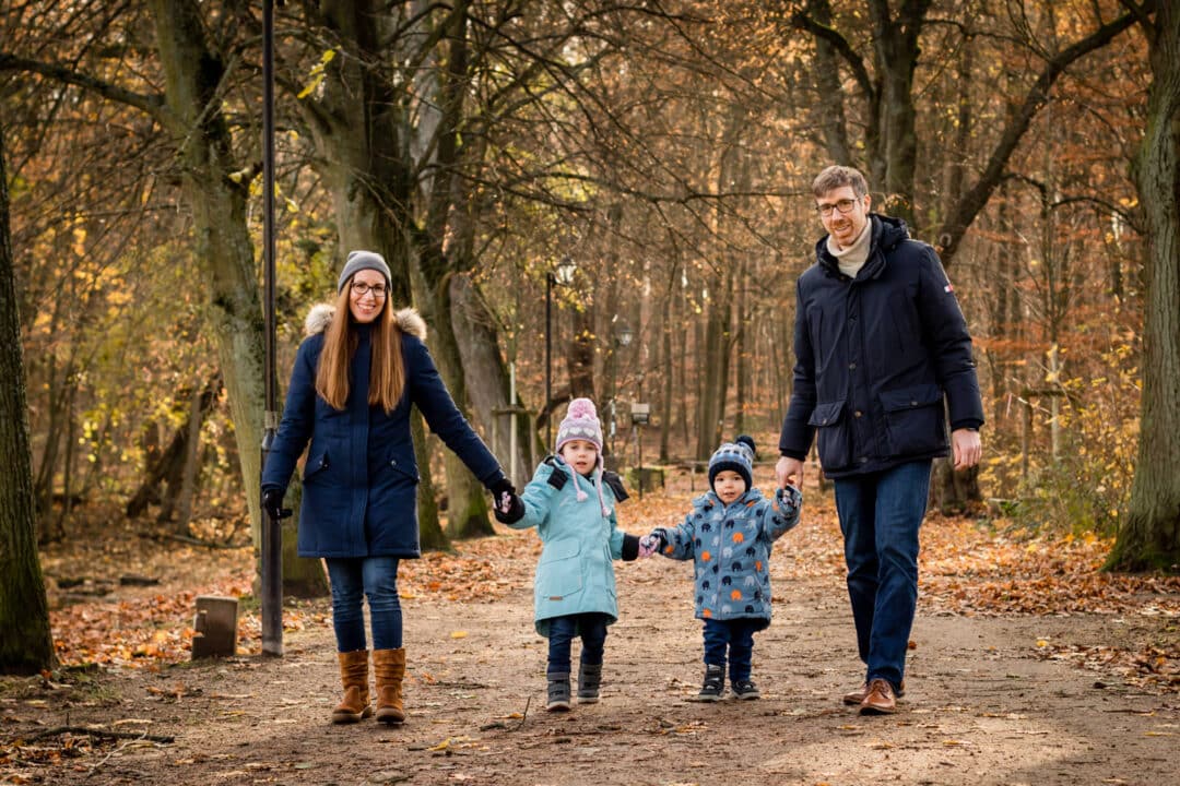 Familie steht zu viert mit Händehalten im Herbstwald und lacht in Kamera