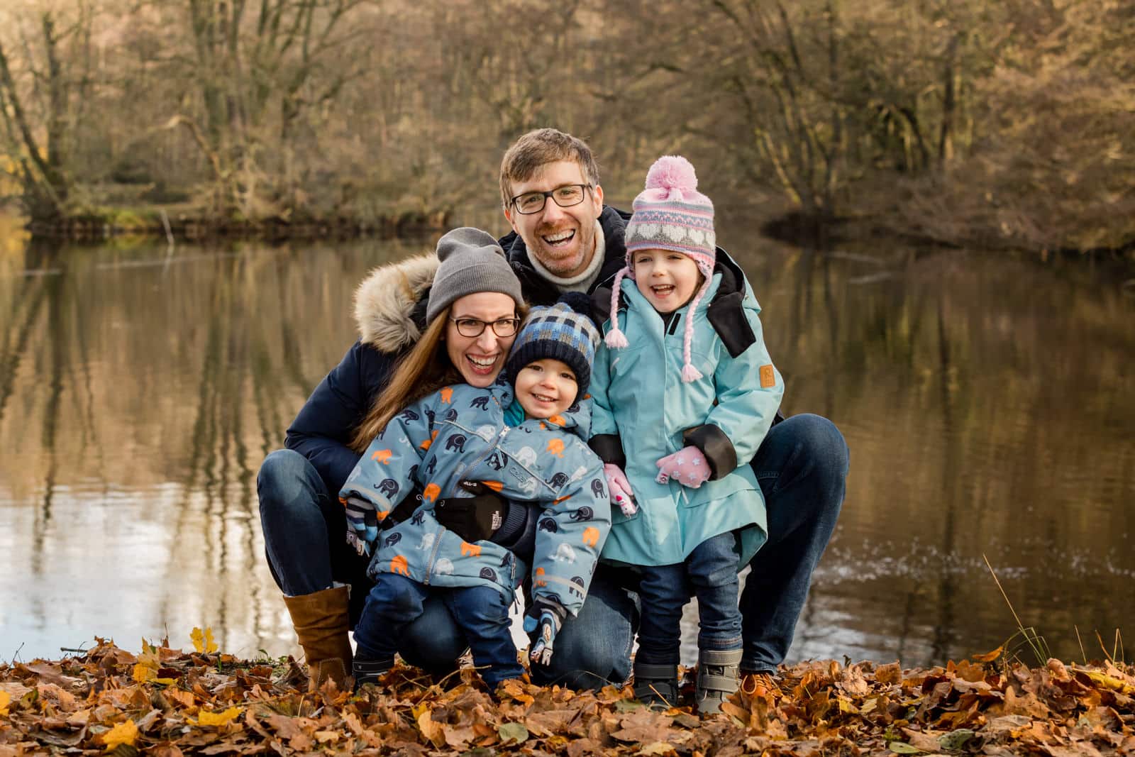 Eine Familie zu viert kniend vor einem Teich mit Herbstlaub