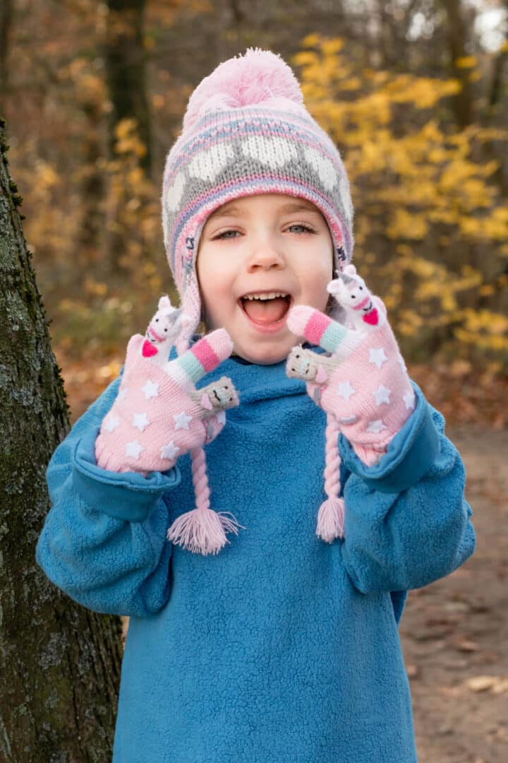 Mädchen mit rosa Muetze und suessen Handschuhen lacht mit Händen in die Kamera im Herbst
