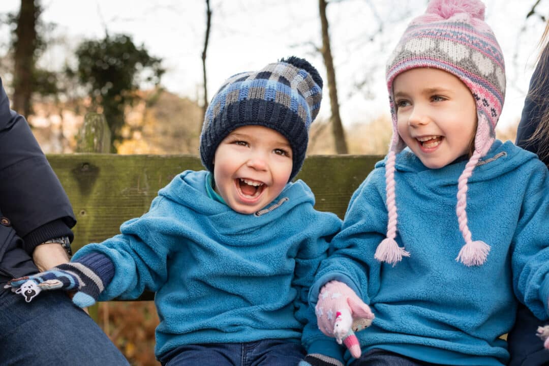 Geschwister sitzen im Herbst mit Mütze auf Bank und lachen