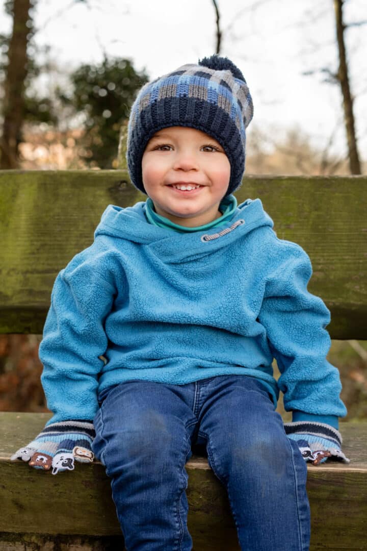 Junge sitzt mit blauer Mütze auf einer Bank im freien und lacht in die Kamera