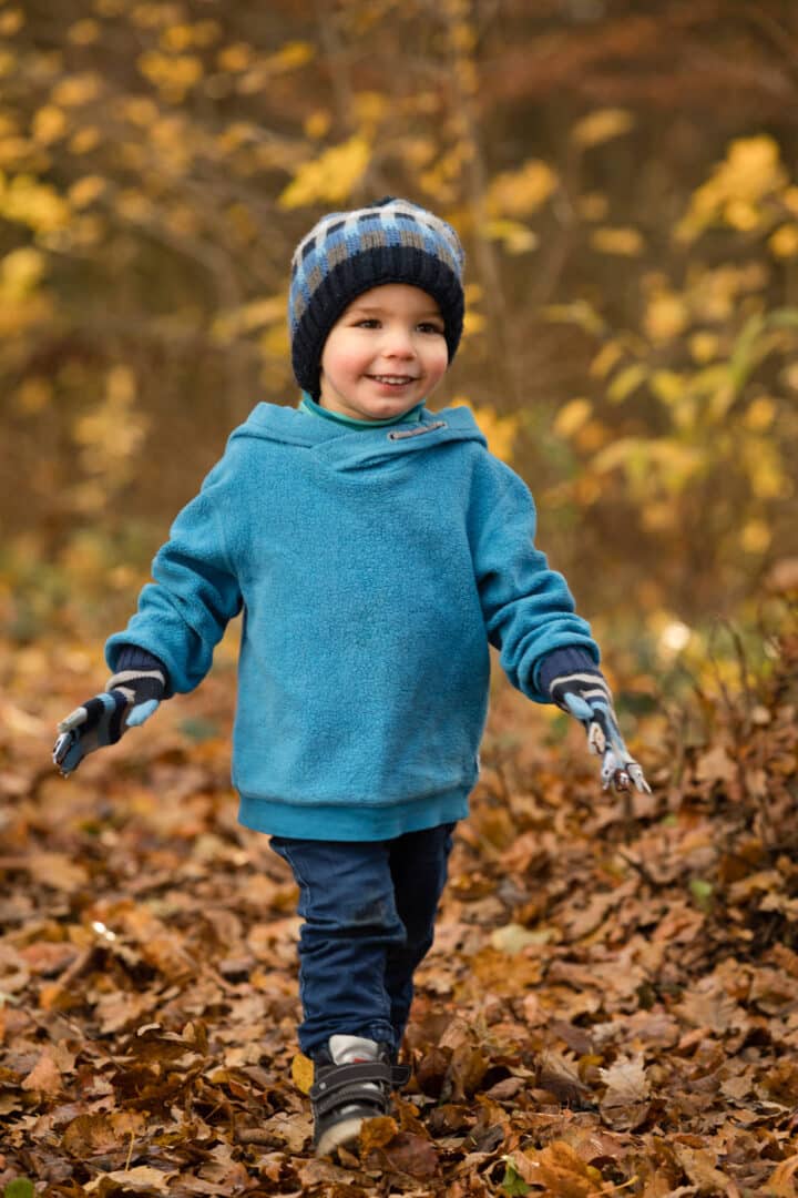 Kleiner lachender Junge läuft im Herbstlaub mit blauem Pullover und Mütze