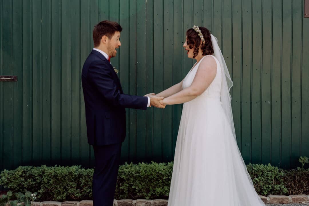 Braut und Bräutigam stehen sich vor dem grünen Hoftor gegenüber, halten sich die Hand und sehen sich beim First Look das erste Mal erstaunt an.