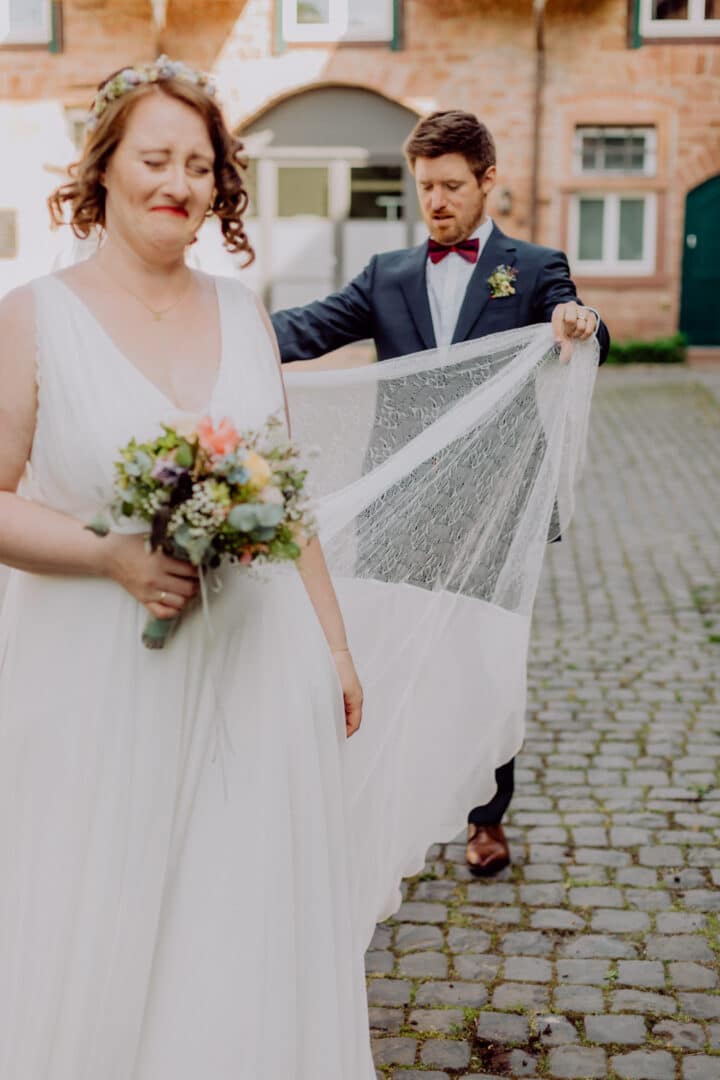 Braut lacht während ihr Mann versucht den Schleier zu sortieren auf dem Hofgut Habitzheim bei ihrer Hochzeit.
