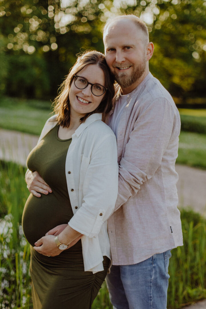 Ein Paar steht zusammen und er und sie halten ihren Babybauch beim Babybauchshooting in einem Darmstädter Garten.