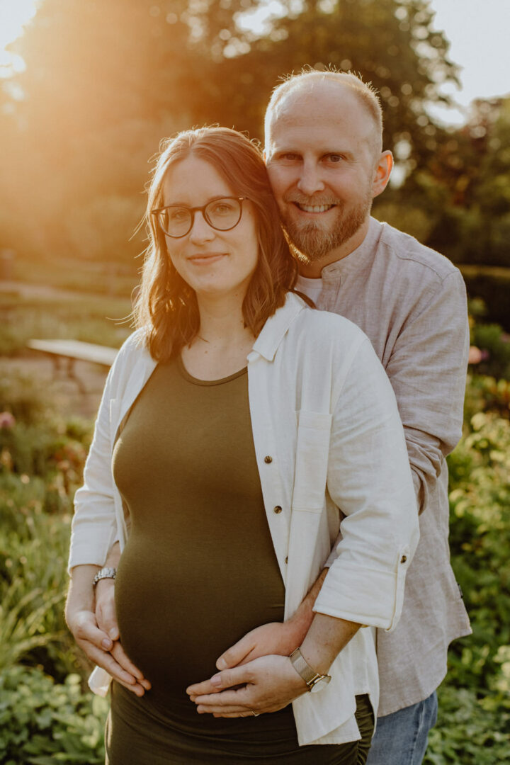 Paar steht im grünen Garten mit Sonnenuntergang und sie hat einen Babybauch