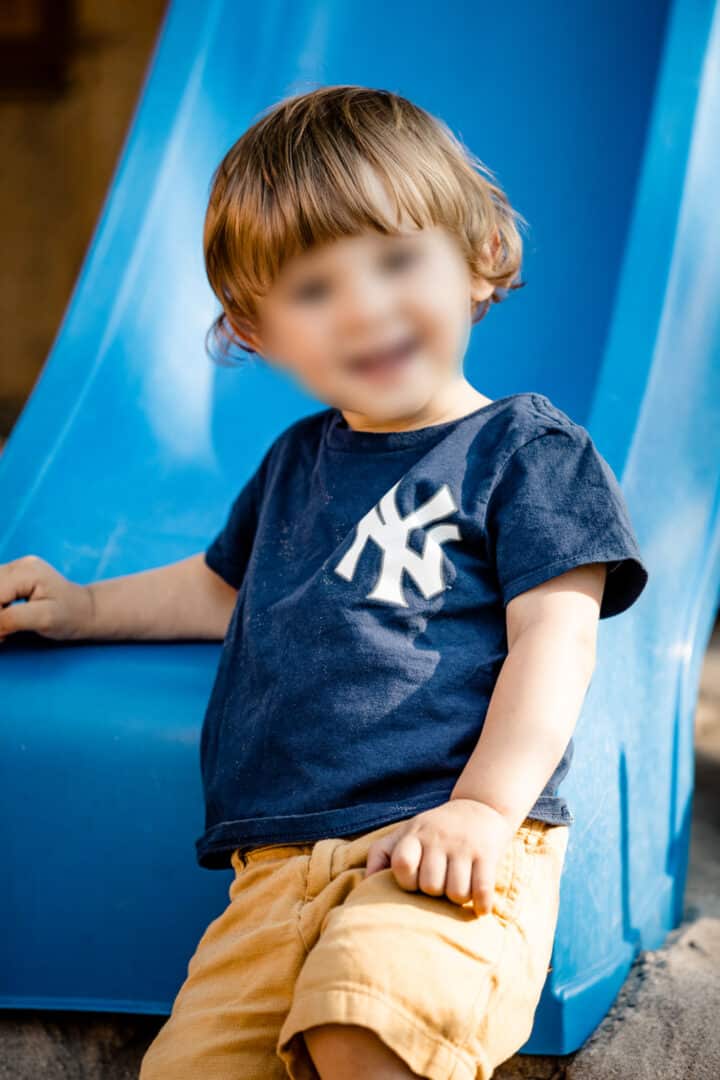 Ein kleiner Junge im blauen T-Shirt lehnt an eine Rutsche und lacht in die Kamera