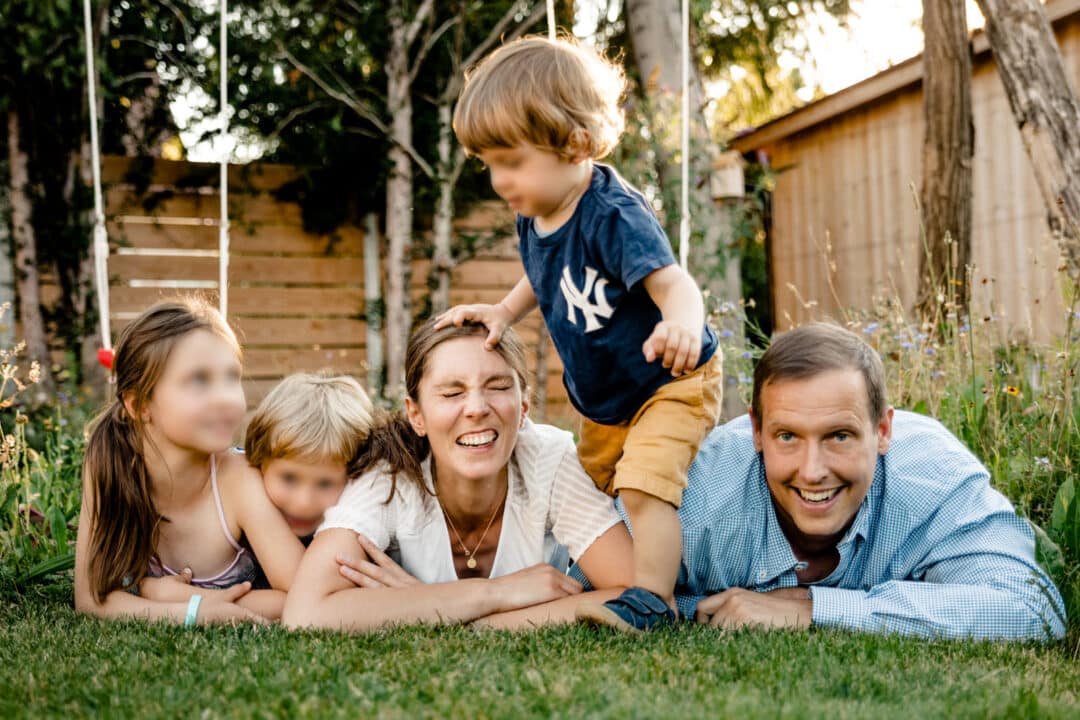 Eine Familie liegt auf dem Rasen und lacht in die Kamera während der Sohn über alle drüber steigt