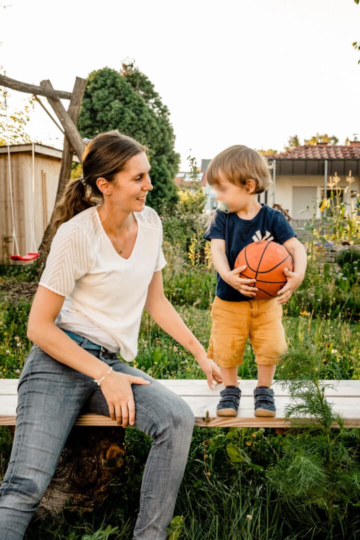 Mutter sitzt auf Bank und neben ihr steht ihr Sohn mit einem Basketball in der Hand und schauen sich an