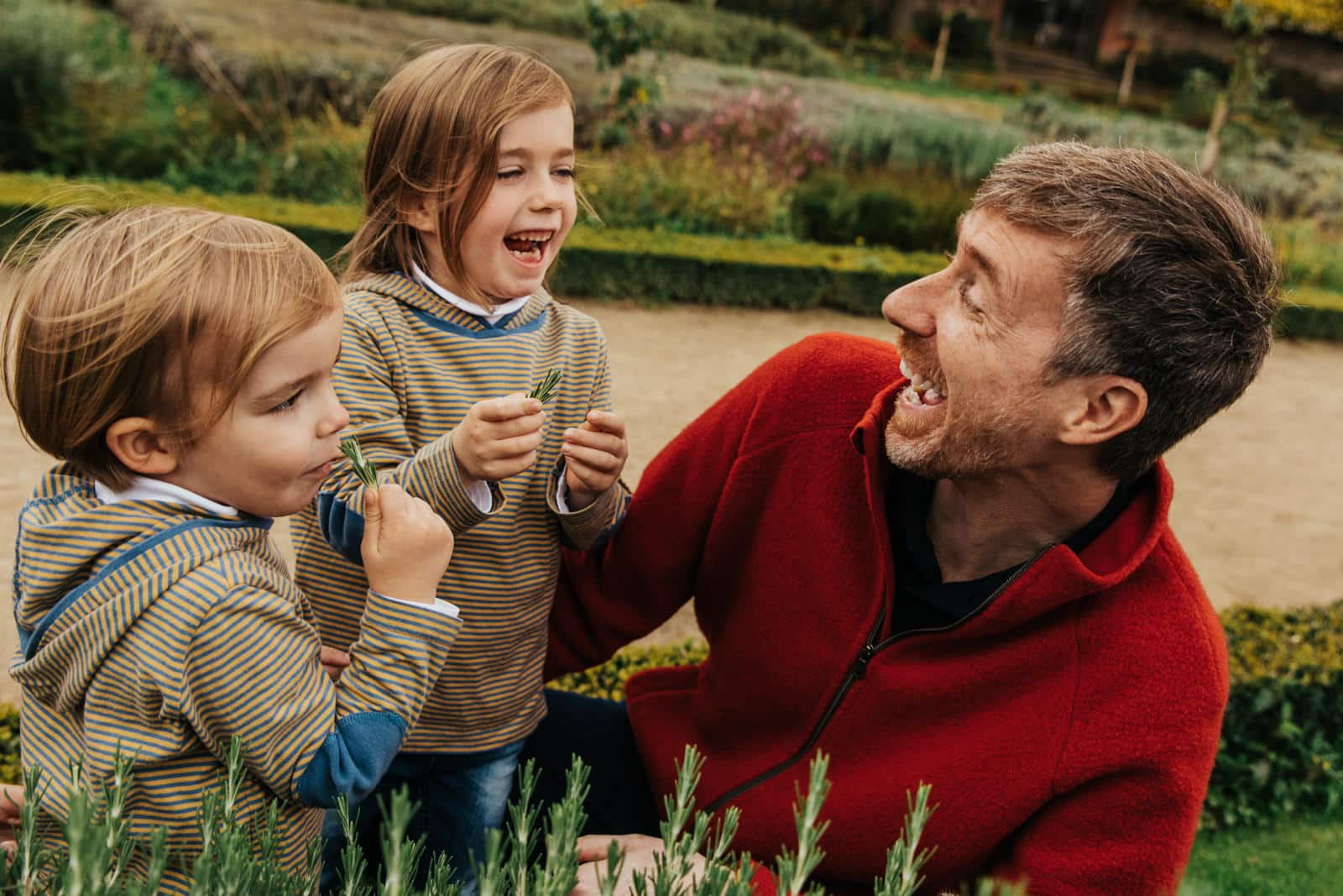 Ein Vater lacht mit seinen zwei Kindern im Park am Rosmarinbusch