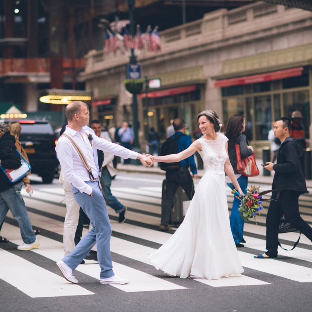 Mann und Frau im Brautkleid auf einem Zebrastreifen in New York City. Halten sich an den Händen und lachen.
