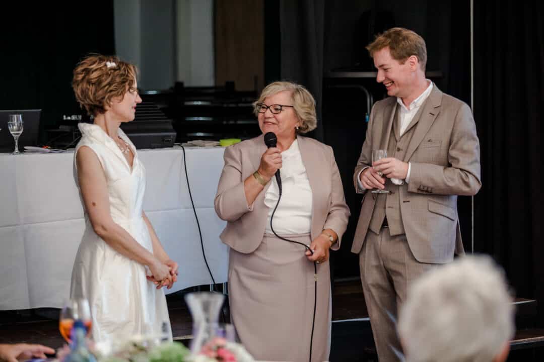 Brautmutter hält Hochzeitsrede zu ihrer Schwiegertochter und Mann lacht mit Glas in Hand im Prinz-Emil-Schlösschen in Darmstadt.