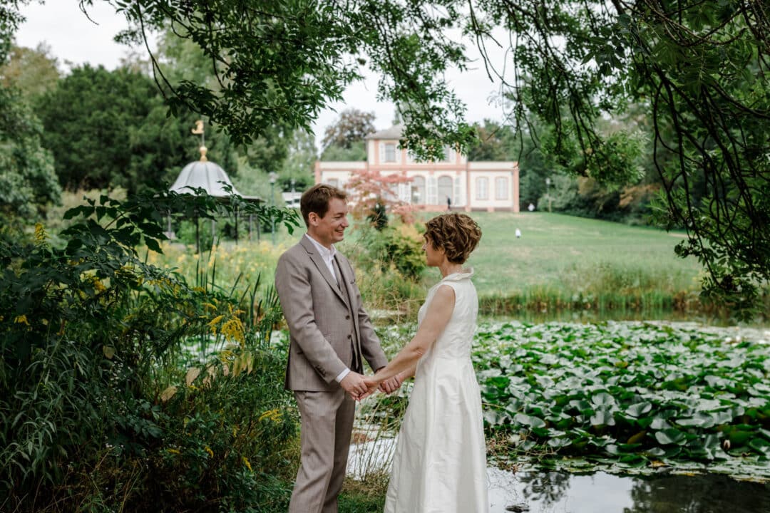 Brautpaar steht Hand in Hand am See im Park des Prinz-Emil-Schlösschen in Darmstadt und lacht sich an.
