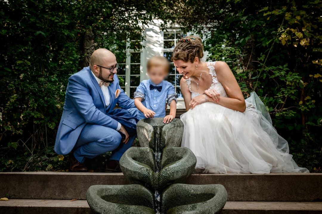 Braut und Bräutigam sitzen vor Wasserfallinstallation und schauen ihrem Sohn zu