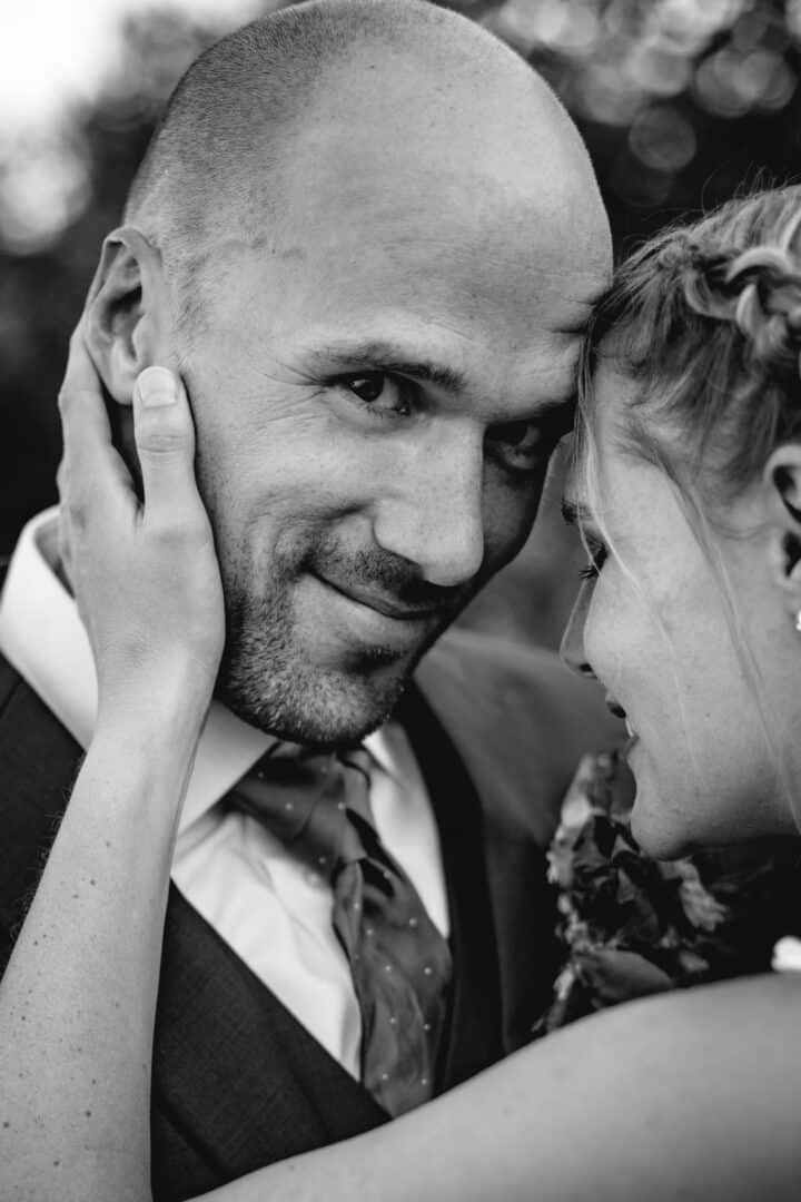 Bräutigam schaut in die Kamera während seine Frau mit ihrer Stirn an seine lehnt und ihre Hand an seinem Hals zärtlich angelegt hat.
