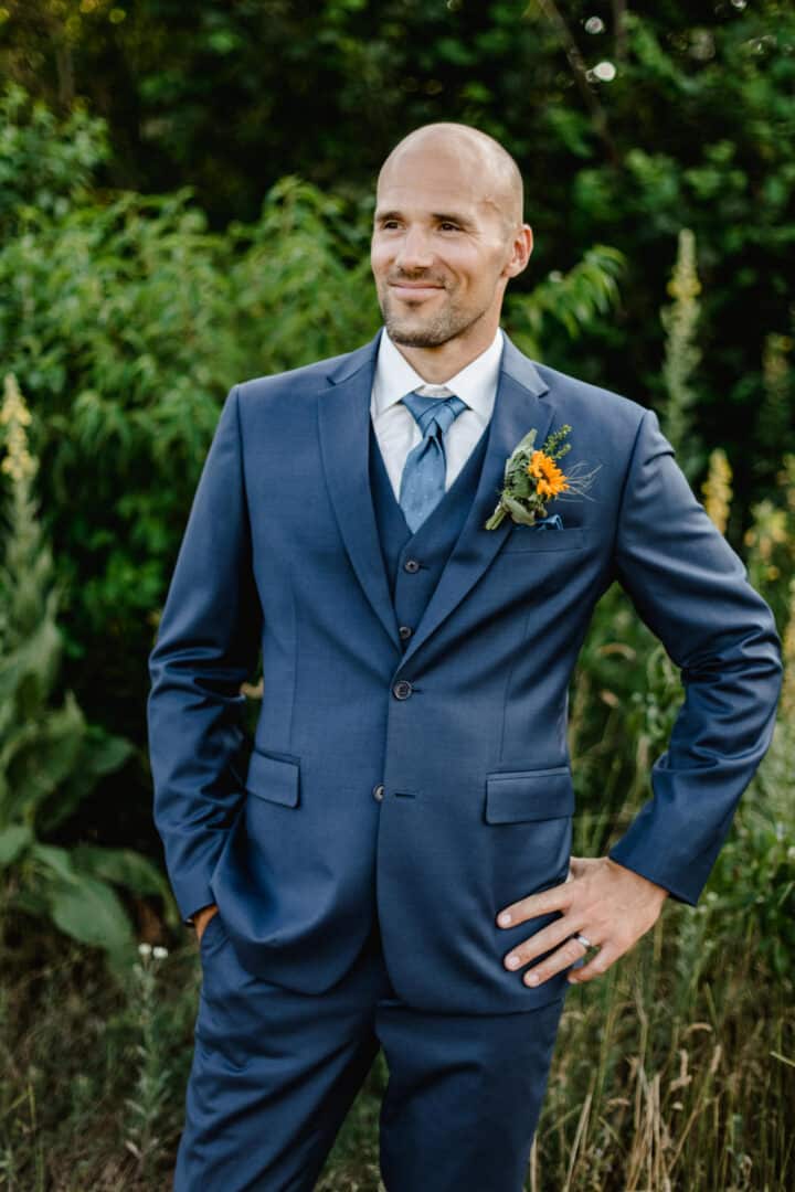 Bräutigam steht im grünen im Hochzeittsanzug mit Hände in den Hüften und lächeld