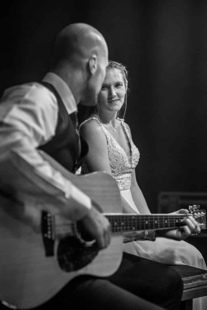 Braut schaut ihren Mann an der auf einer Gitarre auf einer Bühne zu ihr spielt.