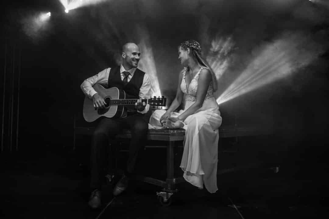 Brautpaar sitzt auf einer Bühne und der Bräutigam spielt Gitarre, Braut hört zu.