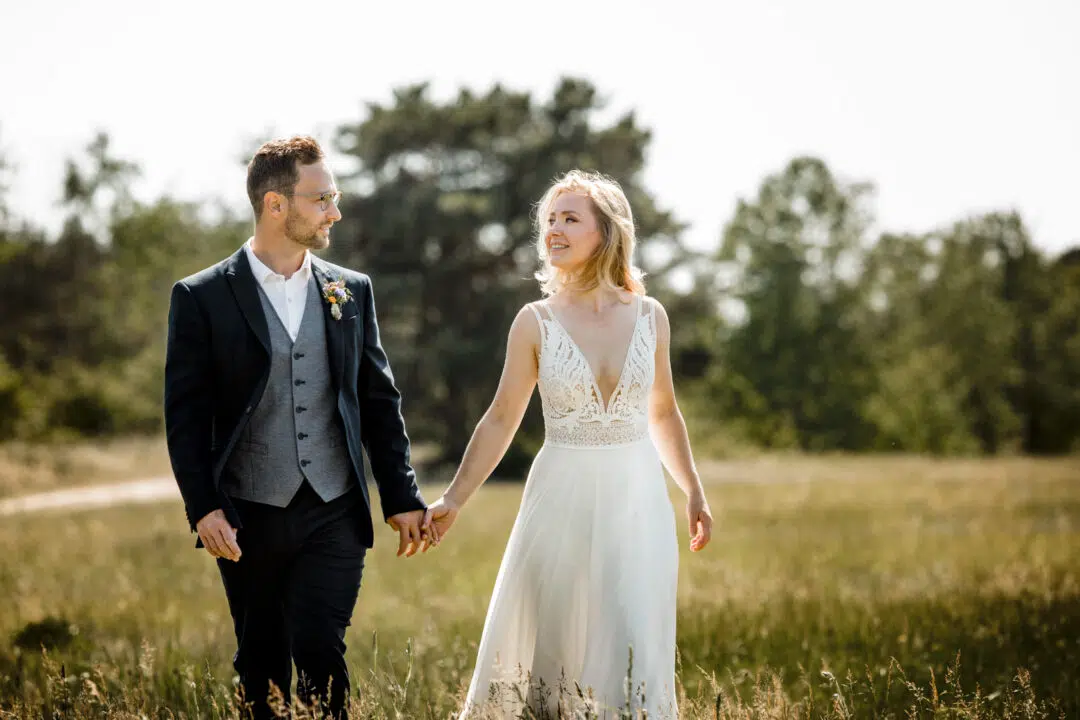 Braut und Bräutigam laufen Hand in Hand über eine Wiese im Sommer bei Sonnenschein und lachen sich an.
