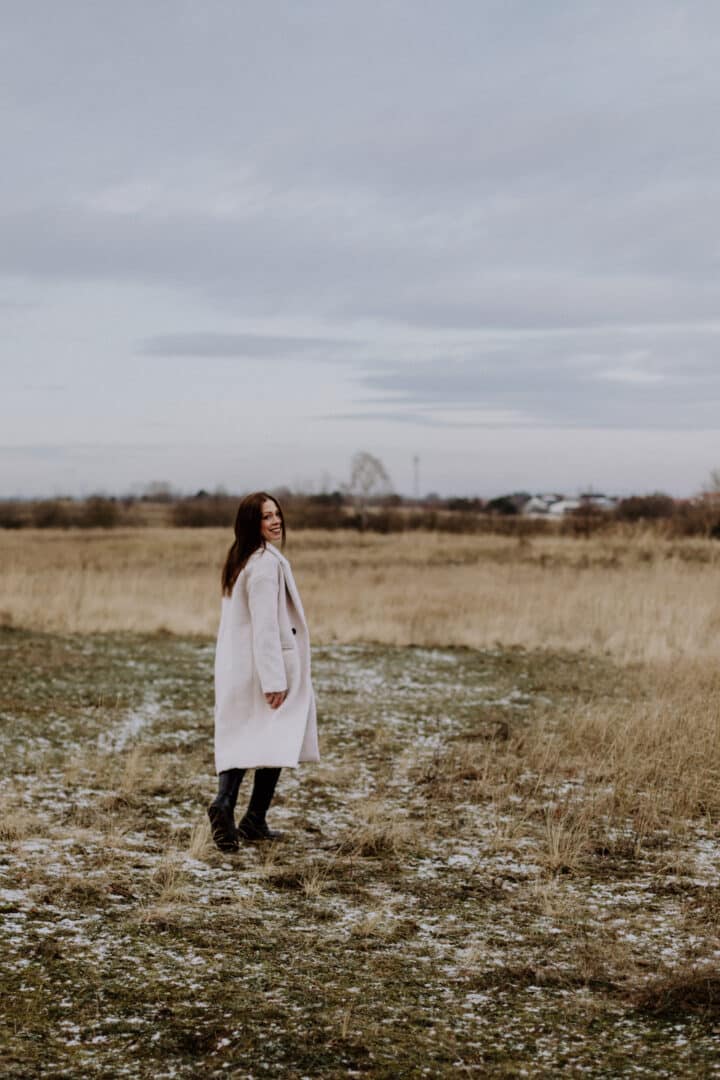 Eine Frau von hinten mit einem weißen Mantel auf einer Wiese mit braunem hohen Gras im Winter.