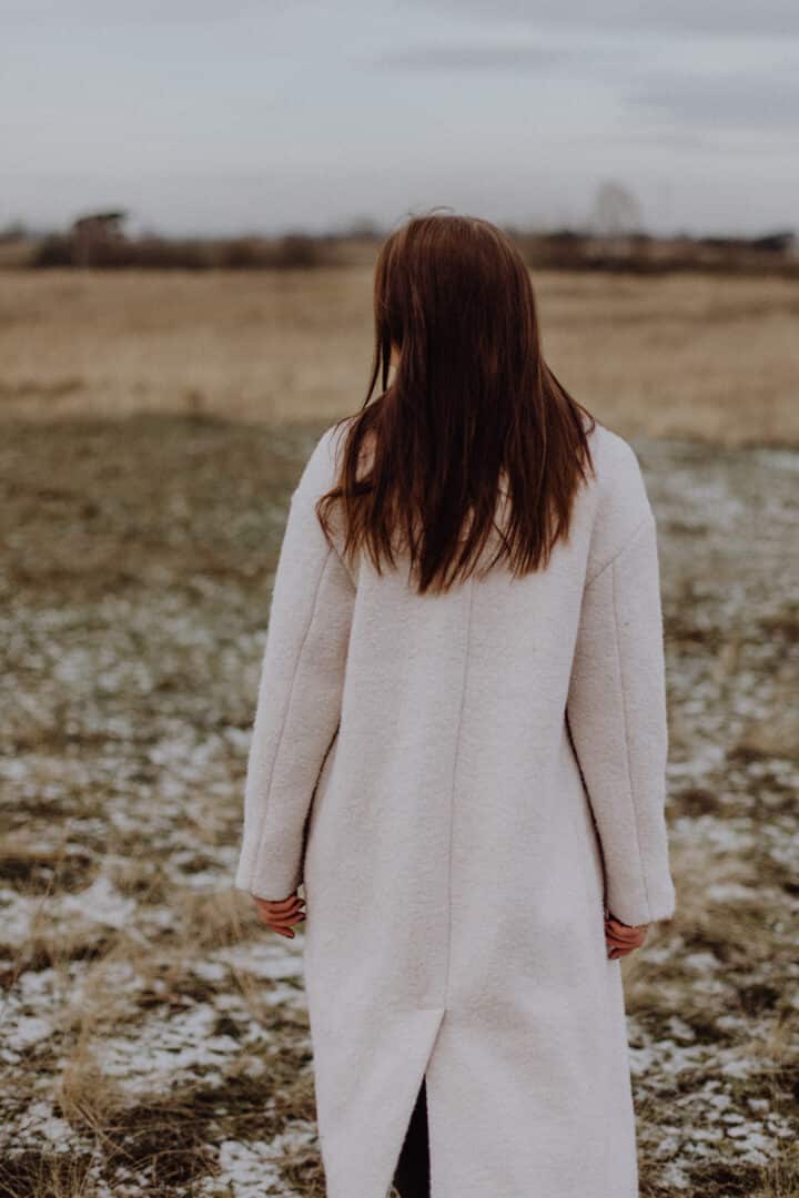 Eine Frau von hinten mit einem weißen Mantel auf einer Wiese mit braunem hohen Gras im Winter.