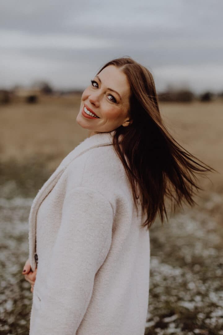 Eine Frau lacht in die Kamera und trägt einen weißen Mantel auf einer Wiese mit braunem hohen Gras im Winter.