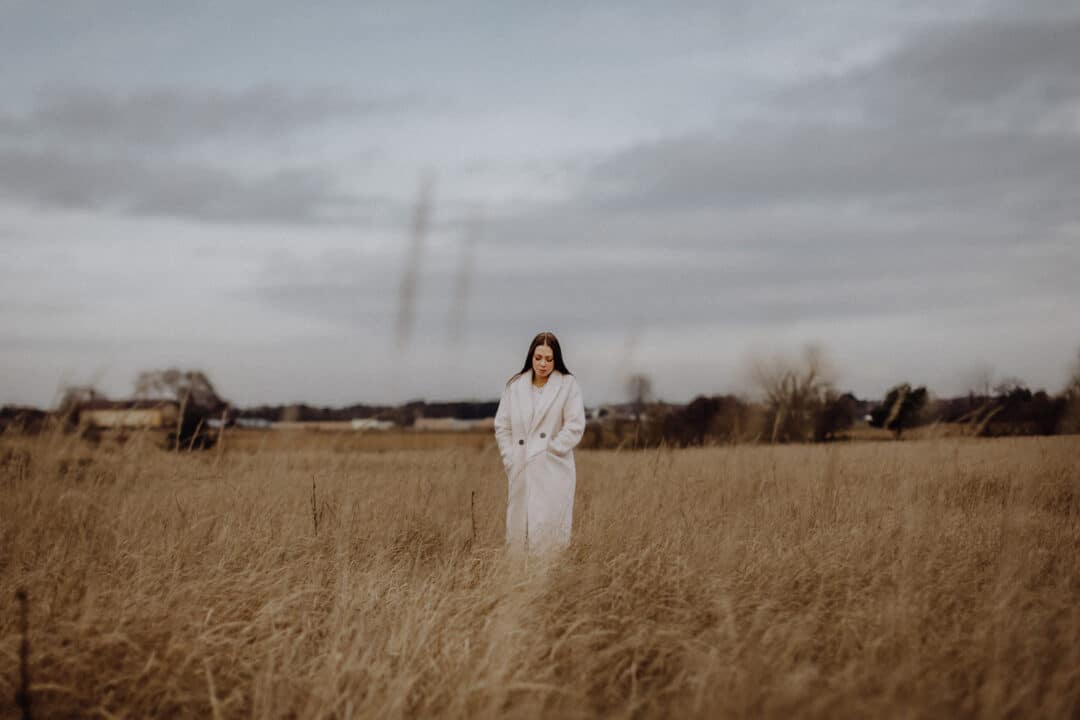 Eine Frau steht mit weißem Mantel auf einer Wiese mit braunem hohen Gras im Winter.