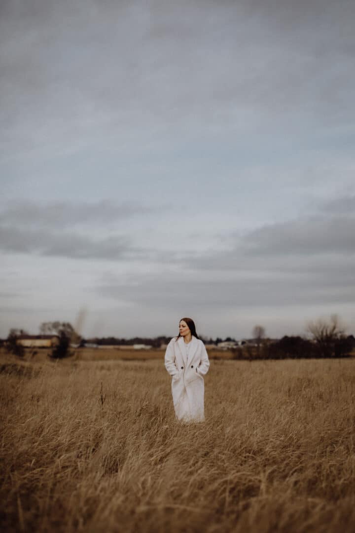 Eine Frau steht mit weißem Mantel auf einer Wiese mit braunem hohen Gras im Winter.