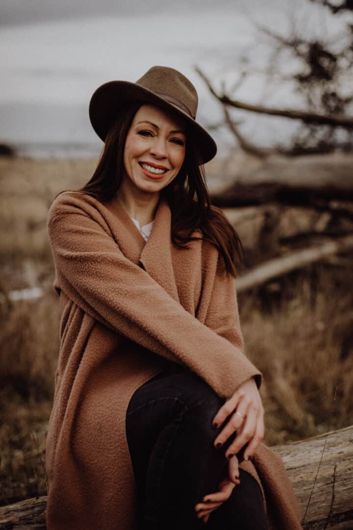 Eine Frau mit dunkelgrünem Hut sitzt in der Natur und lacht in die Kamera.