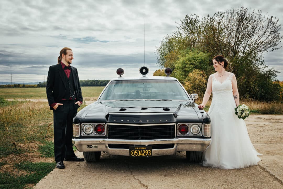 Brautpaar lacht in Kamera und steht vor altem Polizeiauto aus Amerika